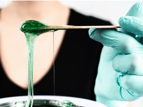 Gloved hand holding a brush with green wax stretching downward over a glass container