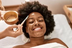 Person with curly hair receiving a facial treatment while lying down, smiling at camera